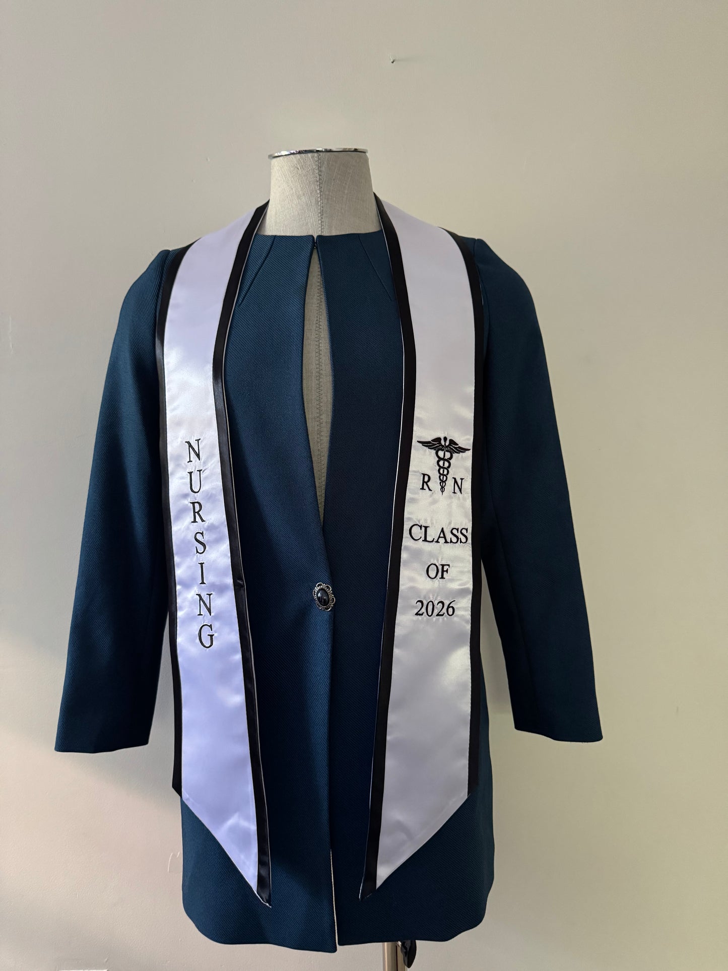 A White With Black Trim RN Graduation Stole, displaying bold vertical 'NURSING' lettering on the right, a medical logo with 'RN' at the left, and 'CLASS OF 2026' embroidered horizontally below. The stole features slanted tips and a white plain background. All characters are embroidered.