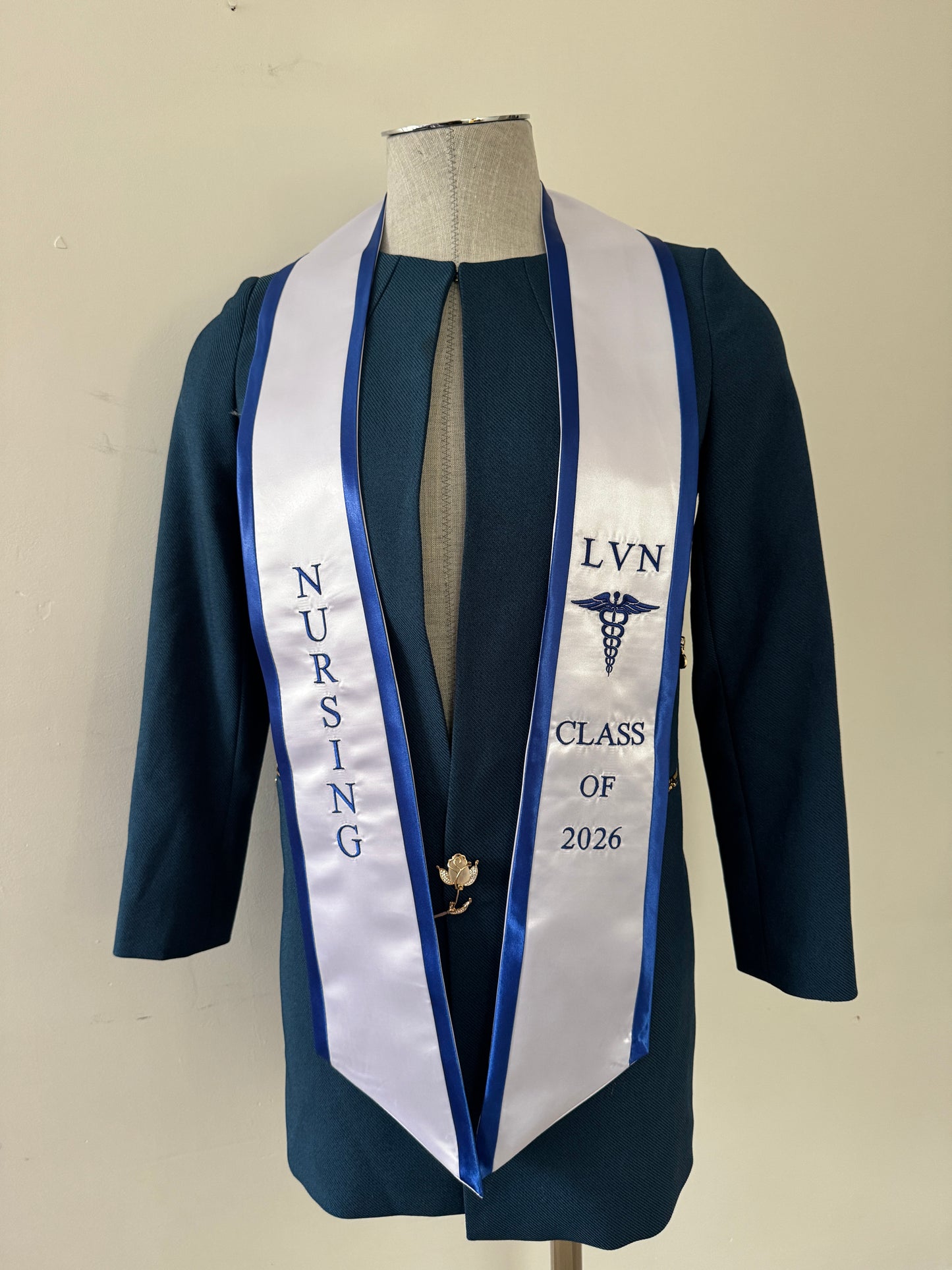 A White With Blue Trim LVN graduation stole, showcasing bold 'LVN' lettering embroidered at the left, followed by a medical logo and 'CLASS OF 2026' embroidered beneath, and 'NURSING' lettering on the right. The stole has slanted tips and a white plain background. All characters are embroidered.