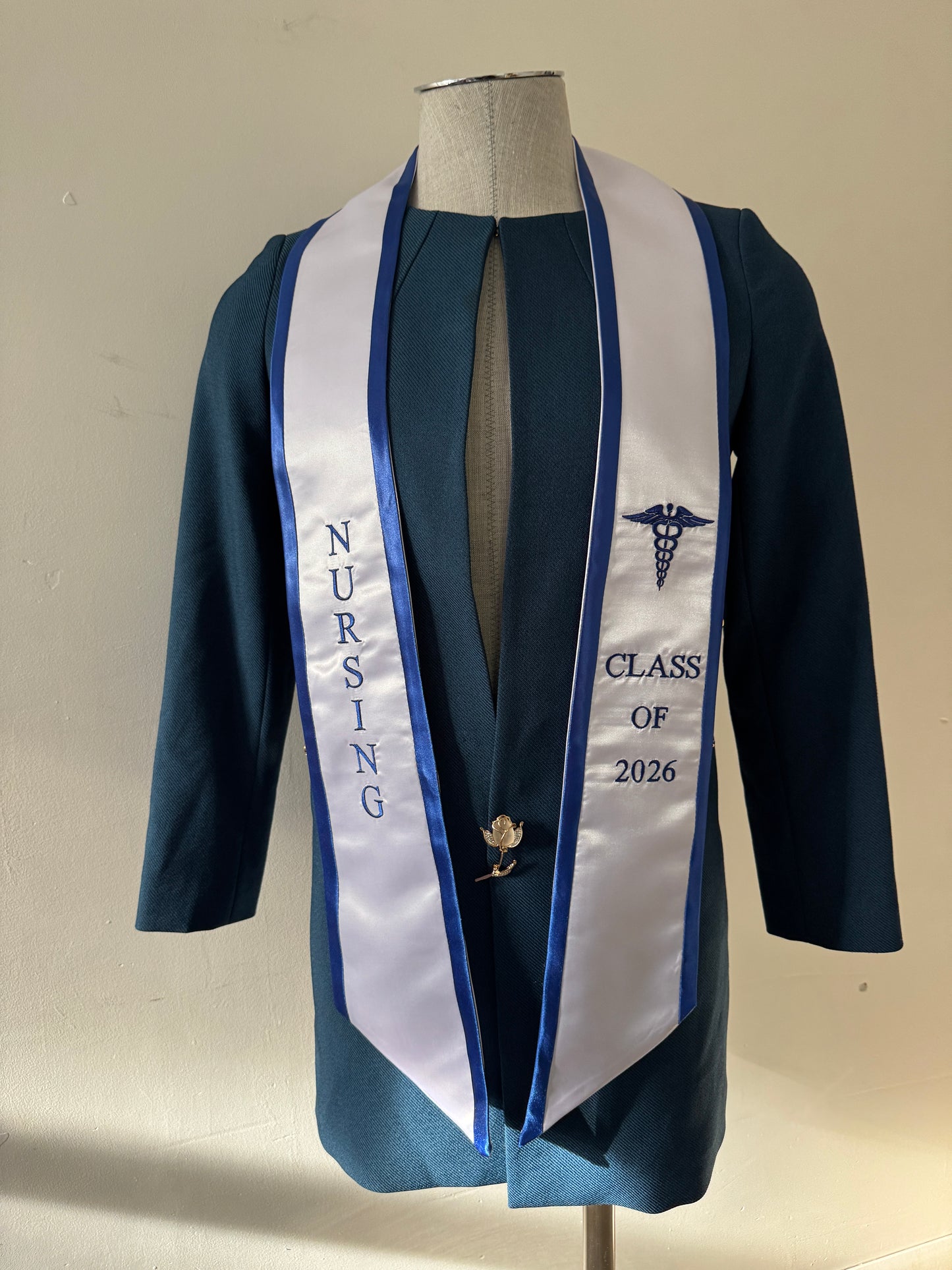 A White With Blue Trim Nursing Graduation Stole, displaying bold 'CLASS OF 2026' lettering embroidered on the left, a medical logo with NURSING on the right embroidered horizontally below. The stole has slanted tips and a white plain background. All text is embroidered.