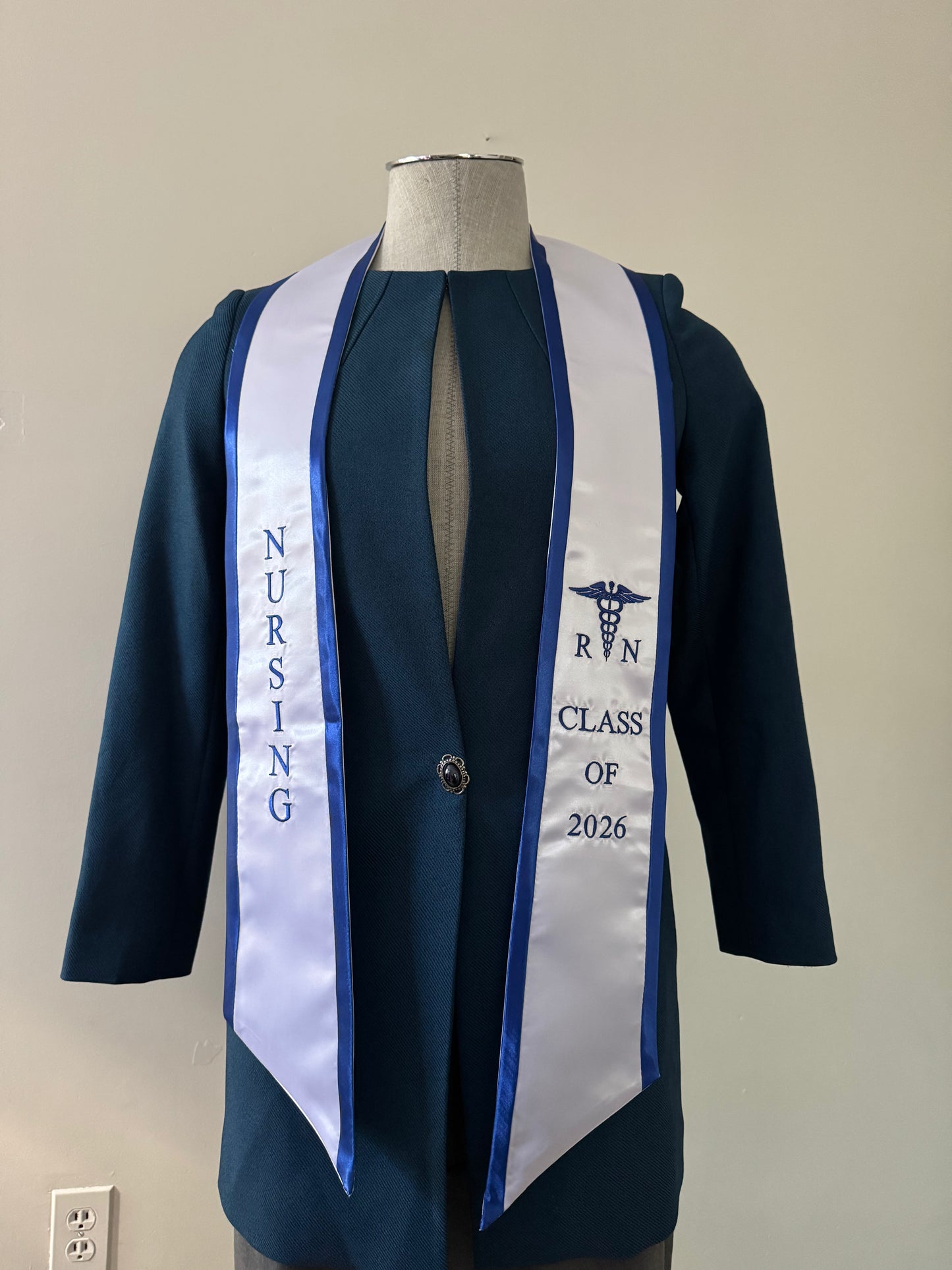 A White With Blue Trim RN Graduation Stole, displaying bold vertical 'NURSING' lettering on the right, a medical logo with 'RN' at the left, and 'CLASS OF 2026' embroidered horizontally below. The stole features slanted tips and a white plain background. All characters are embroidered.