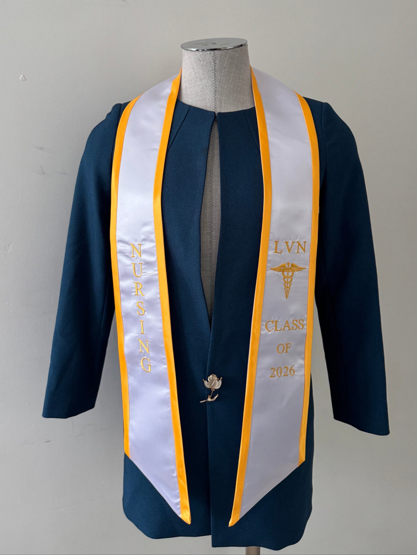 A White With Gold Trim LVN graduation stole, showcasing bold 'LVN' lettering embroidered at the left, followed by a medical logo and 'CLASS OF 2026' embroidered beneath, and 'NURSING' lettering on the right. The stole has slanted tips and a white plain background. All characters are embroidered.