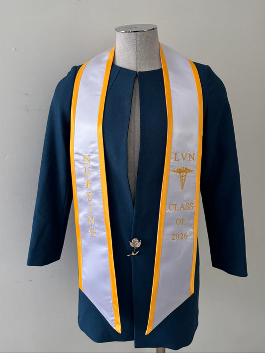 A White With Gold Trim LVN graduation stole, showcasing bold 'LVN' lettering embroidered at the left, followed by a medical logo and 'CLASS OF 2026' embroidered beneath, and 'NURSING' lettering on the right. The stole has slanted tips and a white plain background. All characters are embroidered.