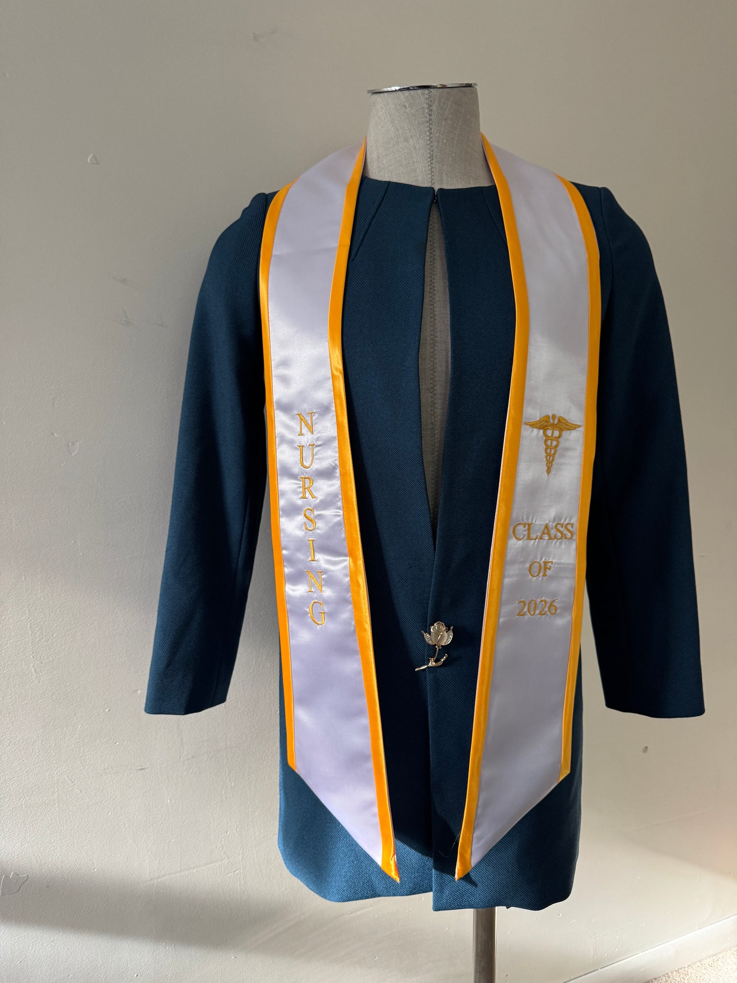 A White With Gold Trim Nursing Graduation Stole, displaying bold 'CLASS OF 2026' lettering embroidered on the left, a medical logo with NURSING on the right embroidered horizontally below. The stole has slanted tips and a white plain background. All text is embroidered.