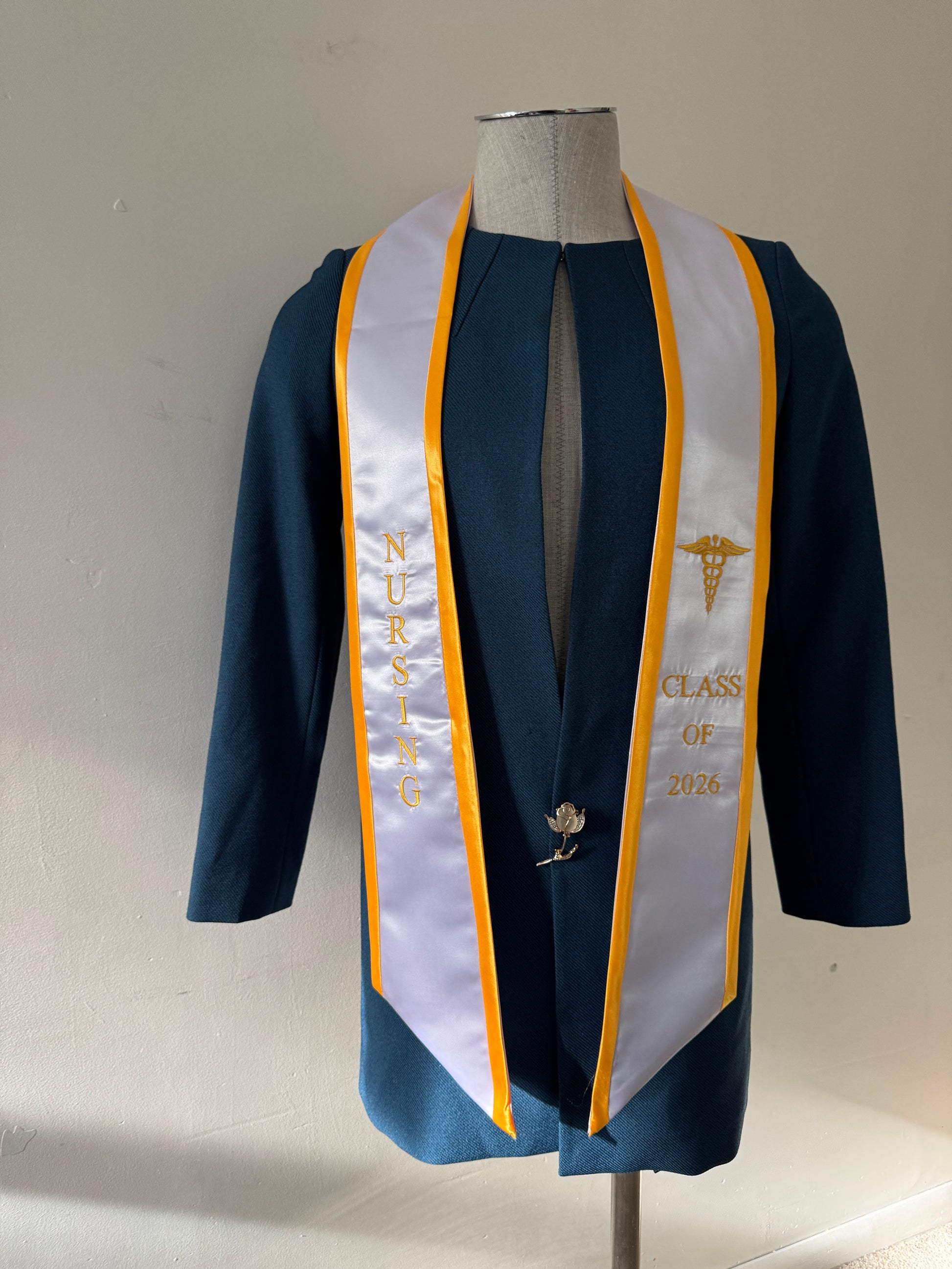 A White With Gold Trim Nursing Graduation Stole, displaying bold 'CLASS OF 2026' lettering embroidered on the left, a medical logo with NURSING on the right embroidered horizontally below. The stole has slanted tips and a white plain background. All text is embroidered.